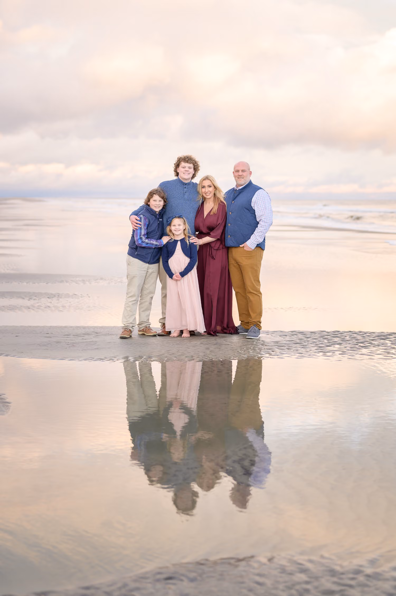 Family of five standing on wet sand with their reflection mirrored below under a pastel sky at Huntington Beach State Park