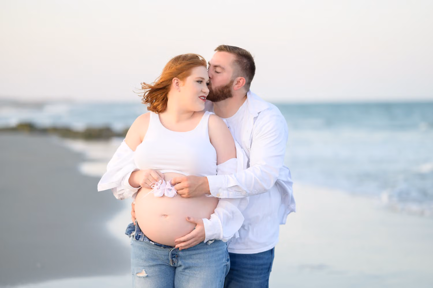 Husband kissing his expectant wife's cheek as they hold tiny baby shoes over her belly by the ocean at The Pelican Inn