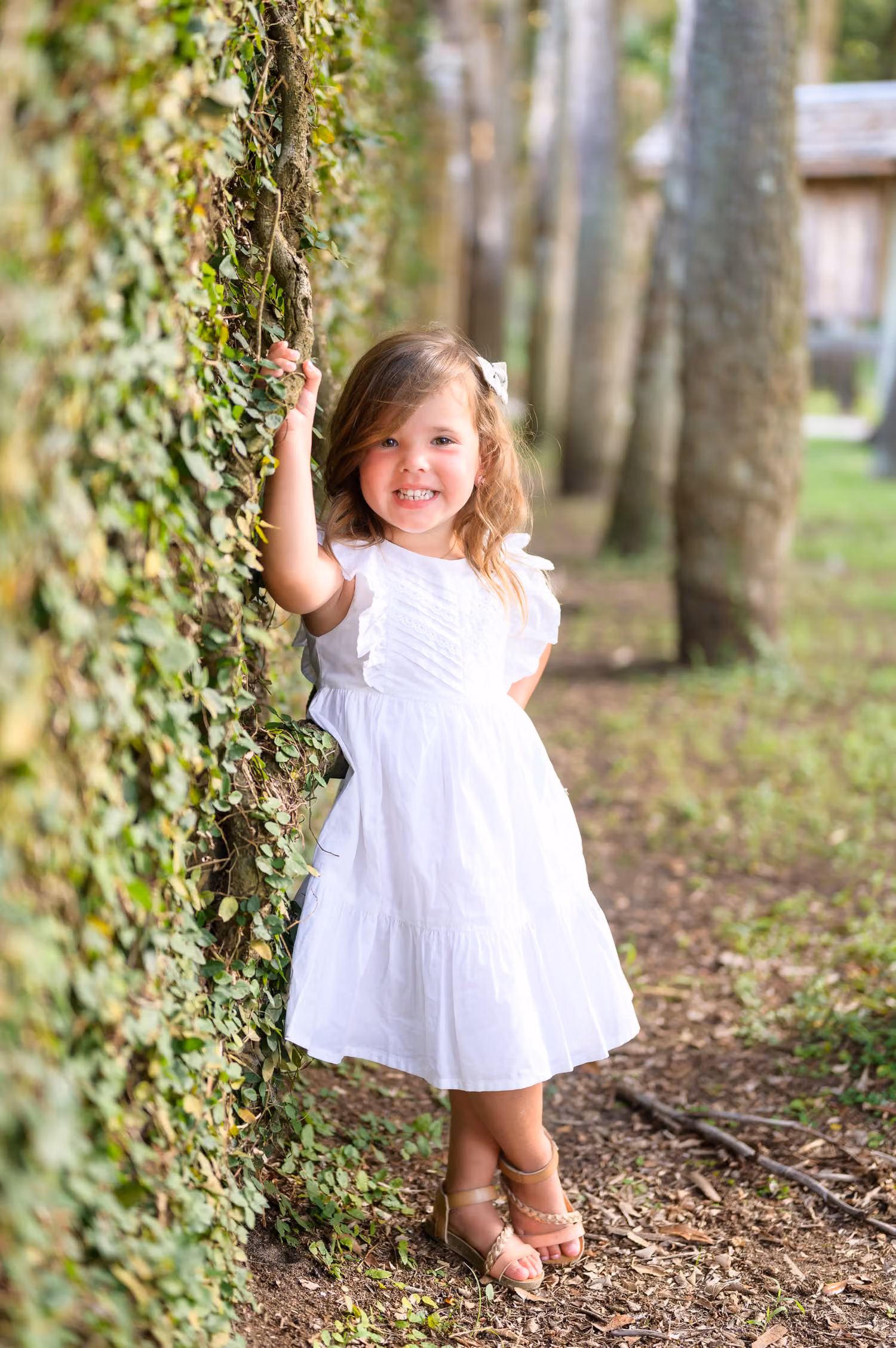 Toddler girl in a white dress grinning as she leans on the ivy wall beside the palms at Huntington Beach State Park