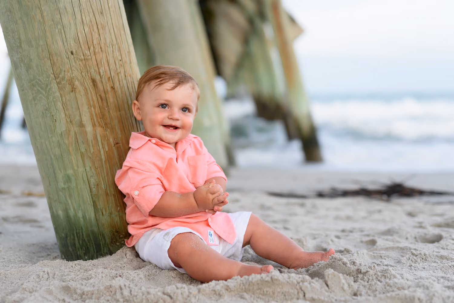 Smiling baby boy in a pink shirt sitting in the sand against a pier piling at Myrtle Beach State Park