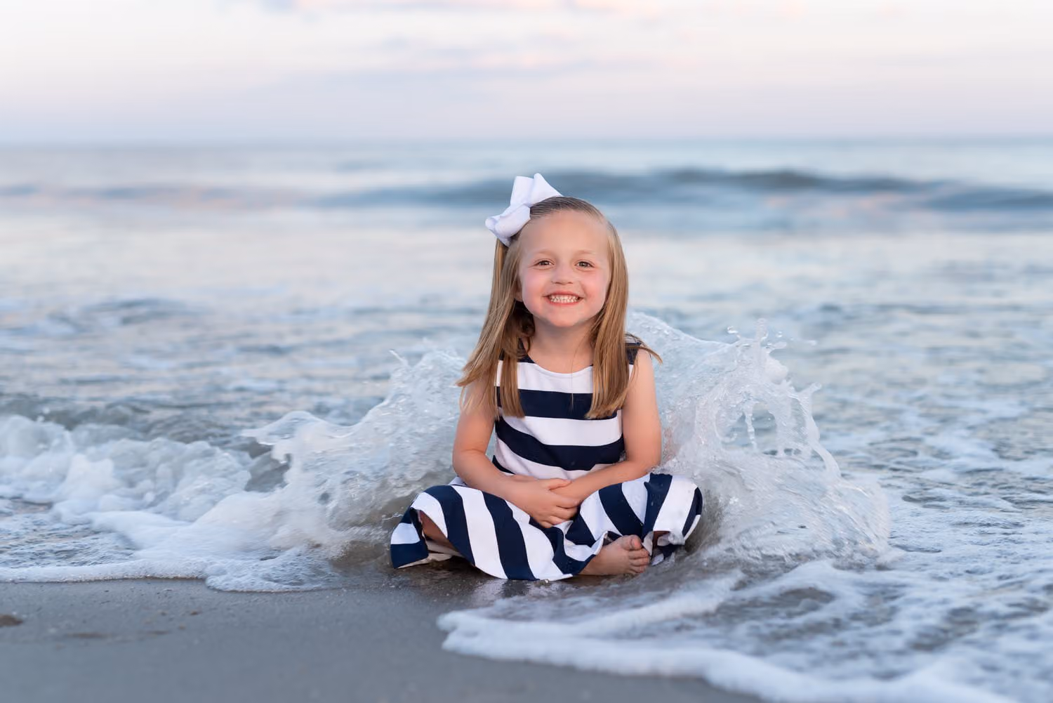 Little girl in a striped dress and hair bow sitting in the surf as a wave splashes around her in Myrtle Beach