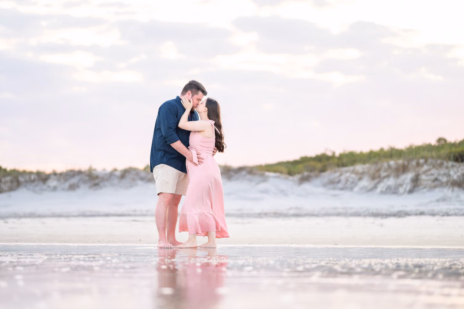 Expectant couple kissing as they stand in shallow water at the ocean's edge, reflected below, at Huntington Beach State Park