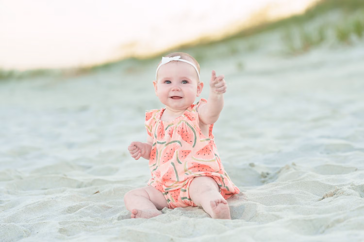 Baby girl in a coral romper and headband sitting in the sand by the dunes giving a thumbs up at Huntington Beach State Park