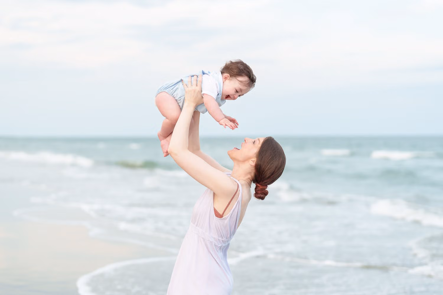 Mom in a pale dress lifting her baby boy above her head in front of the waves at Huntington Beach State Park