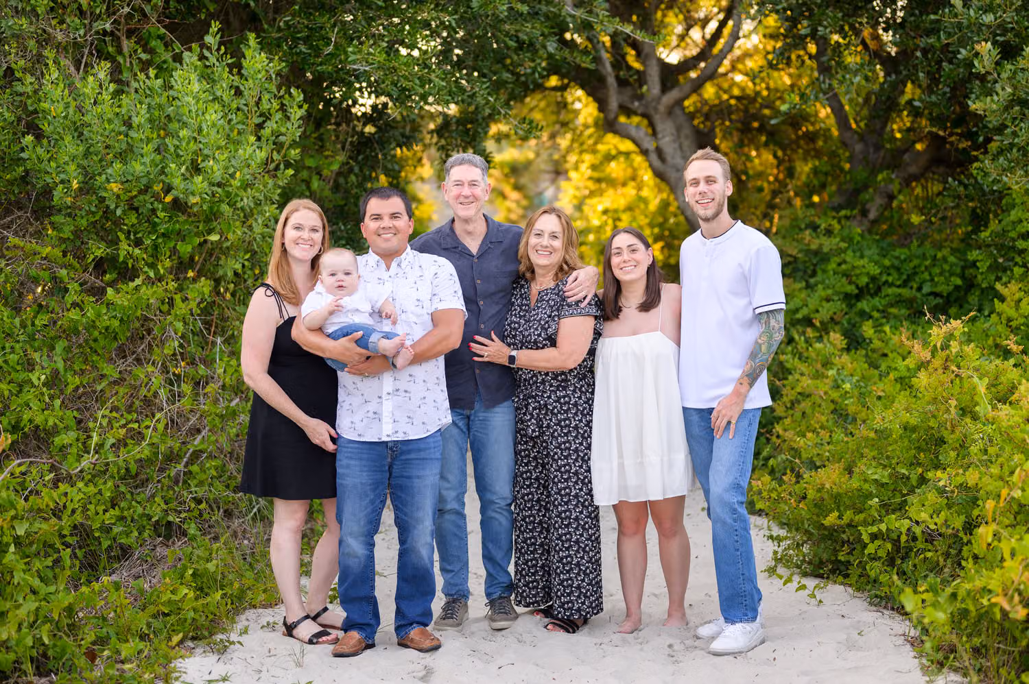 Family of six with a baby boy standing on the sandy path framed by green trees at Huntington Beach State Park