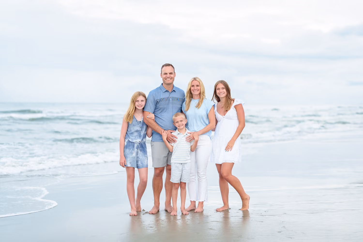 Family of five in blue and white standing by the waves under soft clouds after a rainy day at Huntington Beach State Park