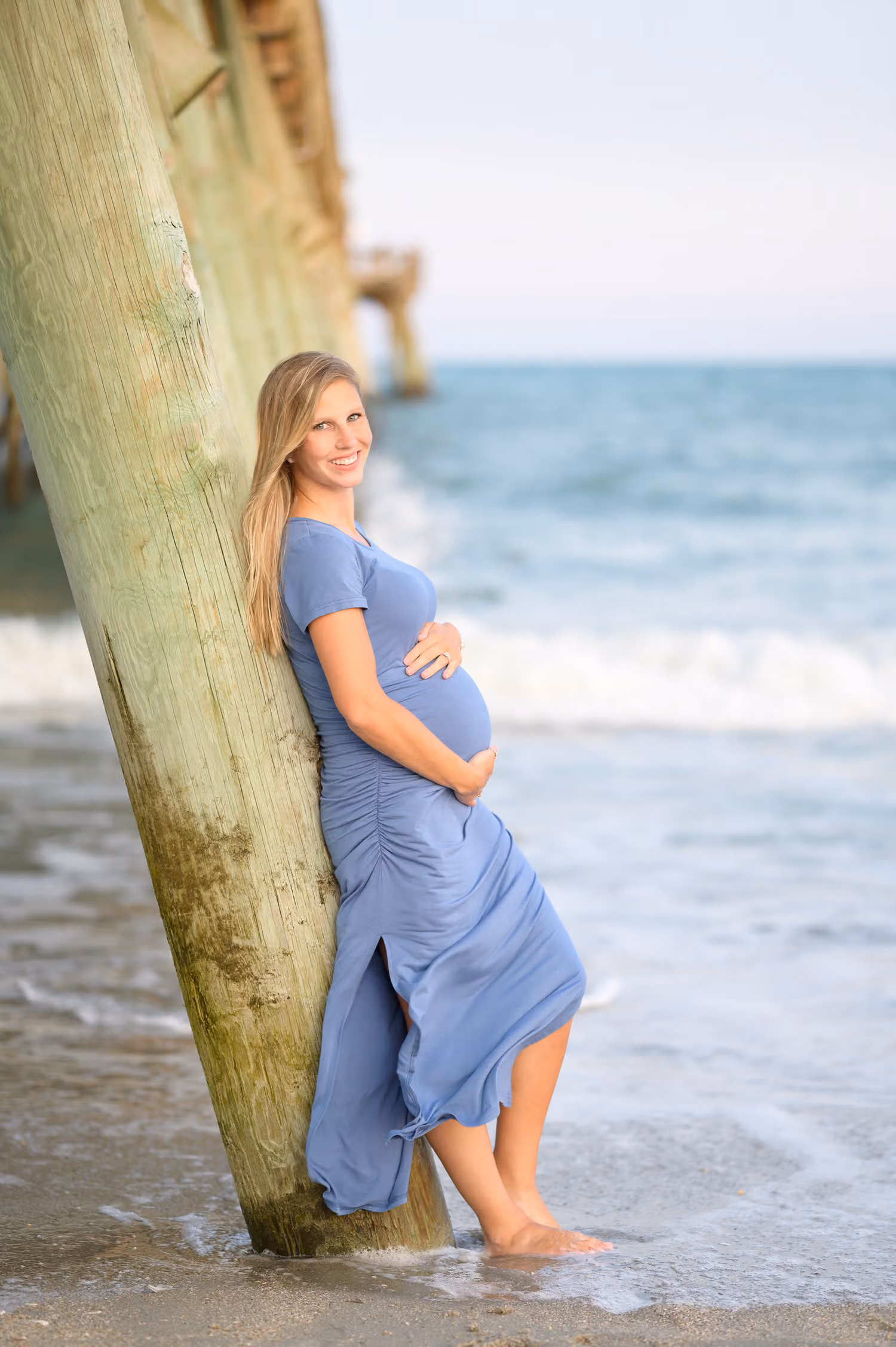 Expectant mother in a blue dress leaning against a pier piling at the water's edge at Myrtle Beach State Park