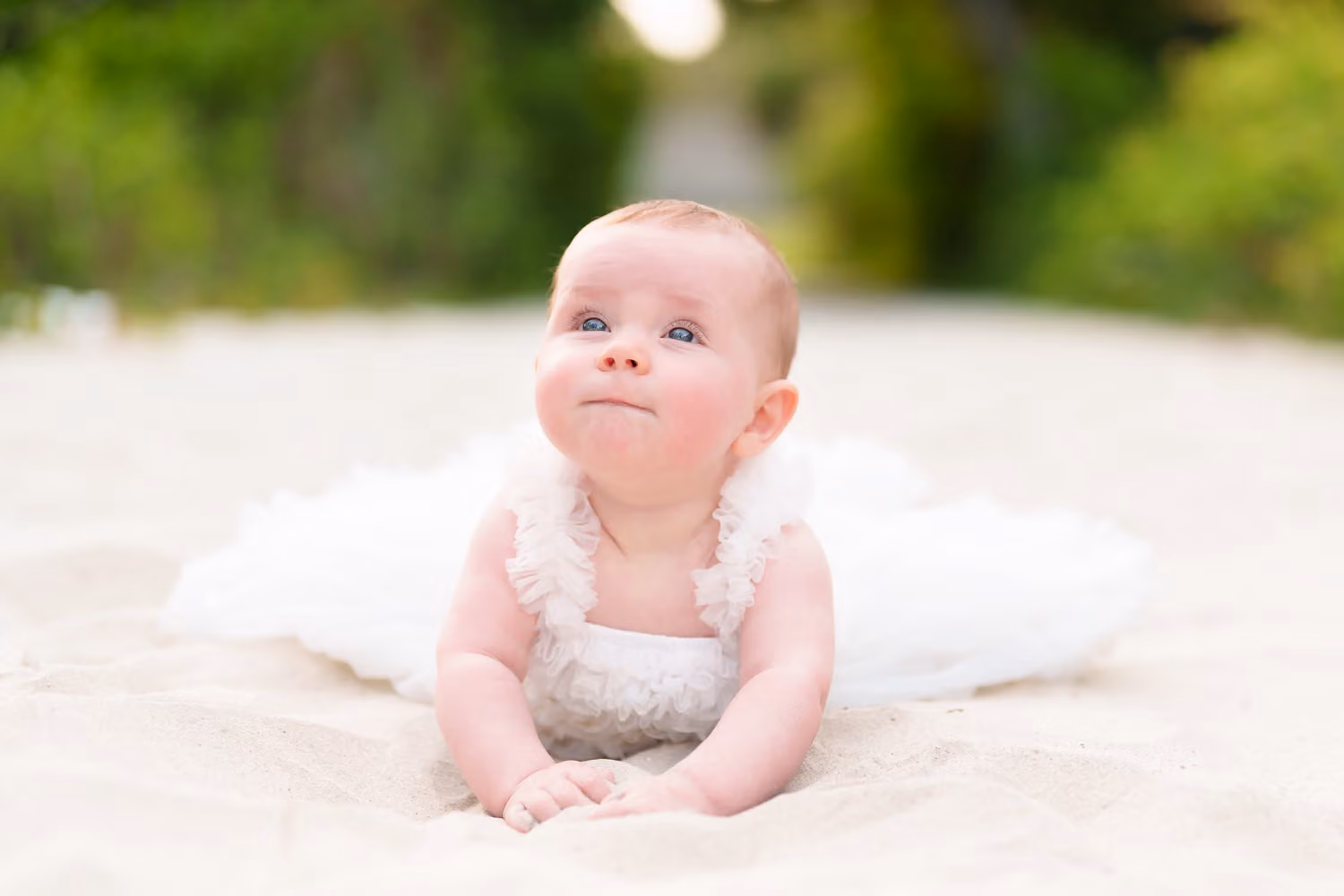 Baby girl in a white tutu lying on her tummy in the sand and gazing upward at Huntington Beach State Park