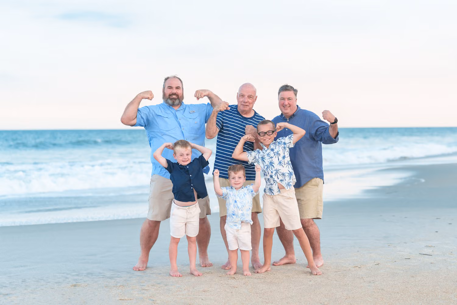 Dads and their little boys flexing their muscles together on the beach in front of the ocean at Huntington Beach State Park