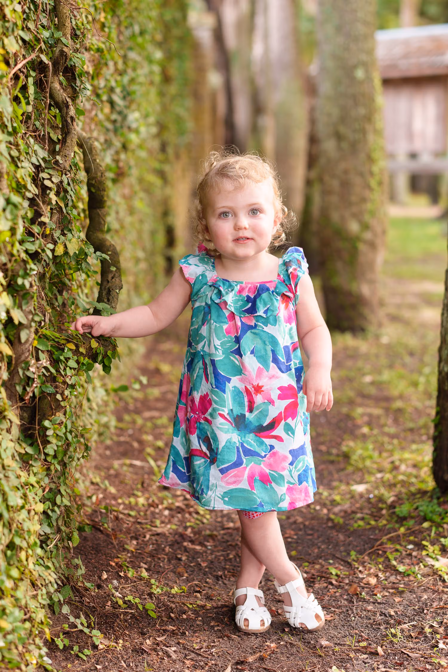 Toddler girl in a floral dress posing by the ivy wall with a sparkle in her eyes at Huntington Beach State Park
