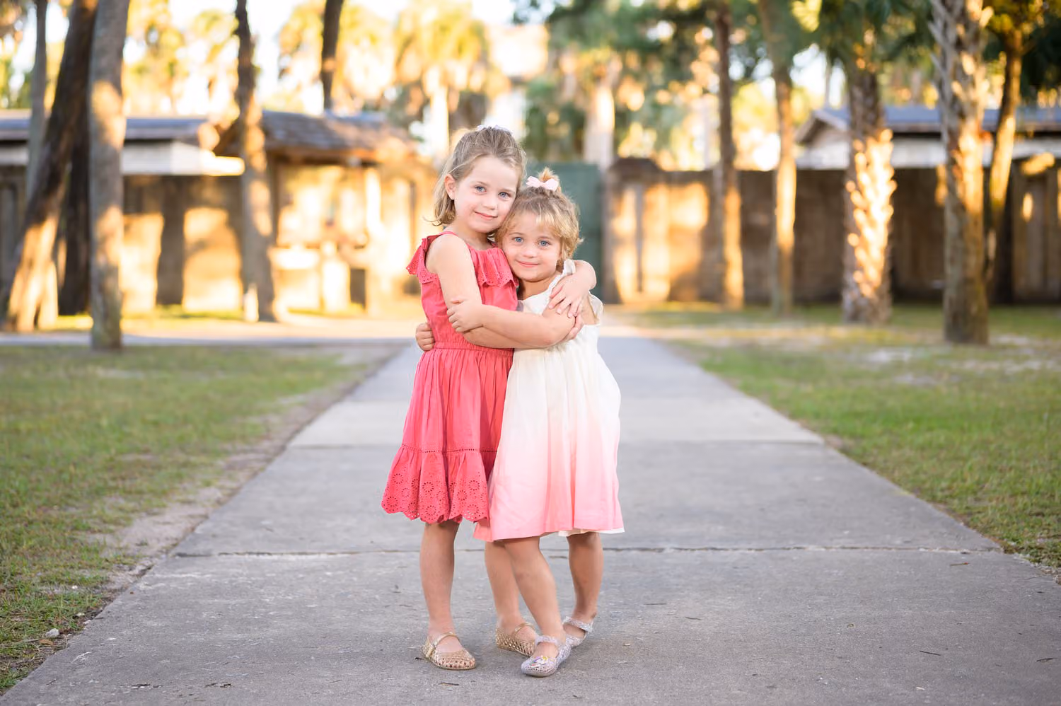 Little sisters in pink dresses hugging on a sidewalk lined with palm trees at Huntington Beach State Park