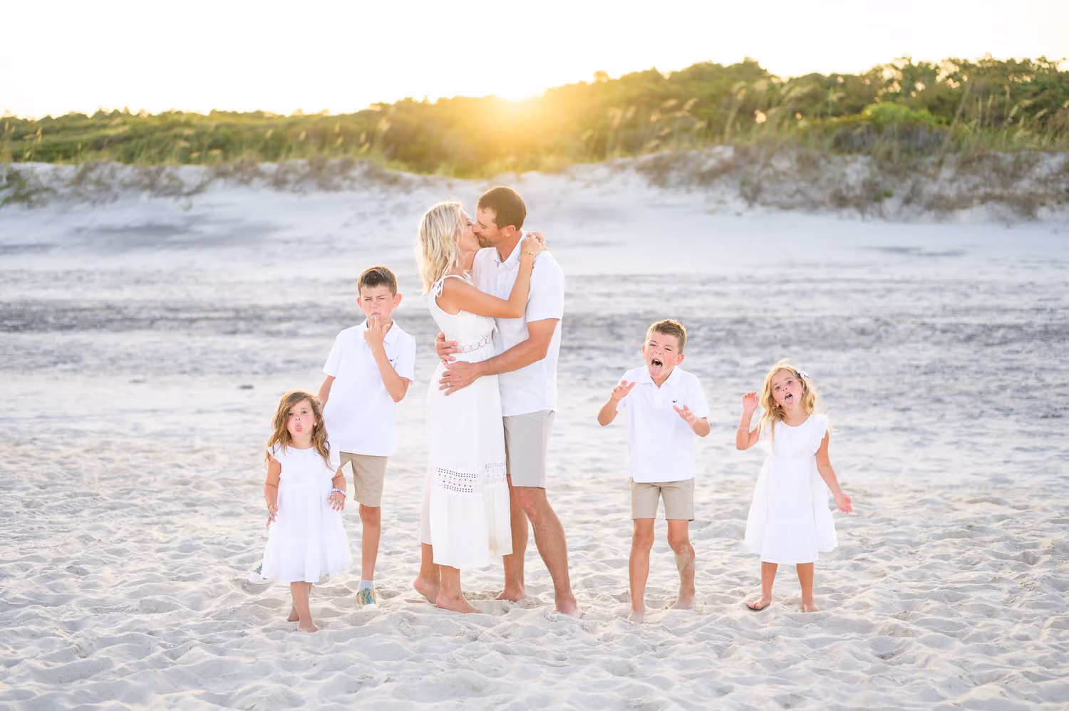 Mom and dad kissing on the beach at sunset while their four kids make silly faces around them at Huntington Beach State Park