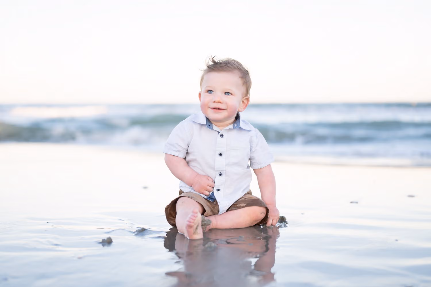 Toddler boy in a button-up shirt sitting on wet sand with his reflection below him at Garden City Beach