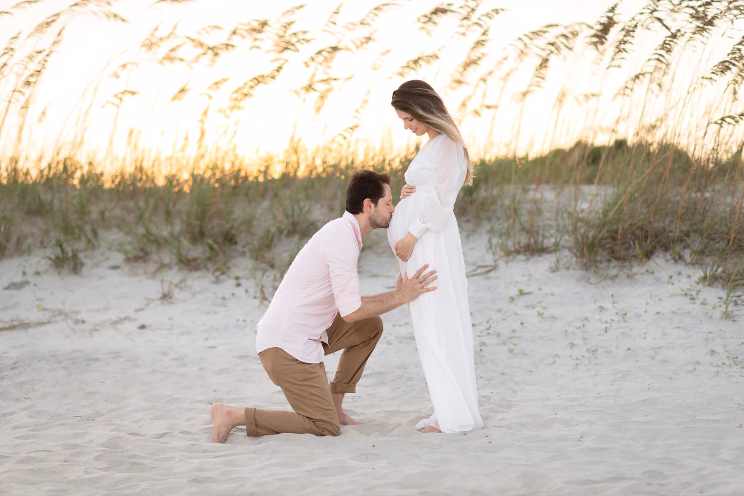Husband kneeling in the sand to kiss his pregnant wife's belly in front of the sea oats at Huntington Beach State Park