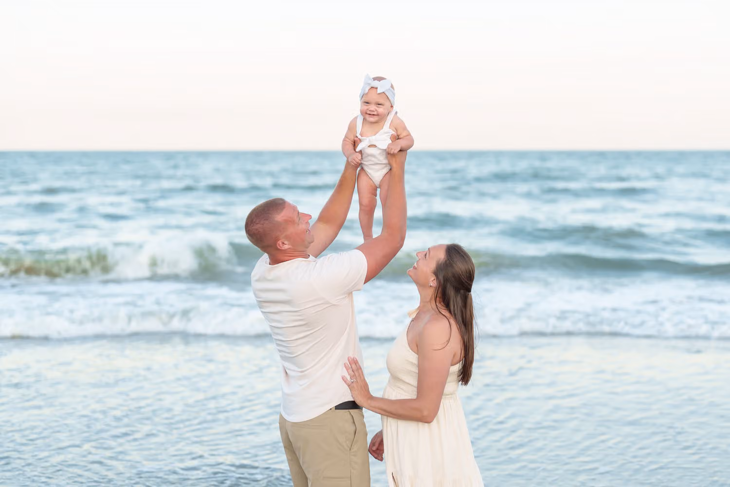 Dad lifting his baby high above the waves while mom looks up and smiles on the beach in Myrtle Beach