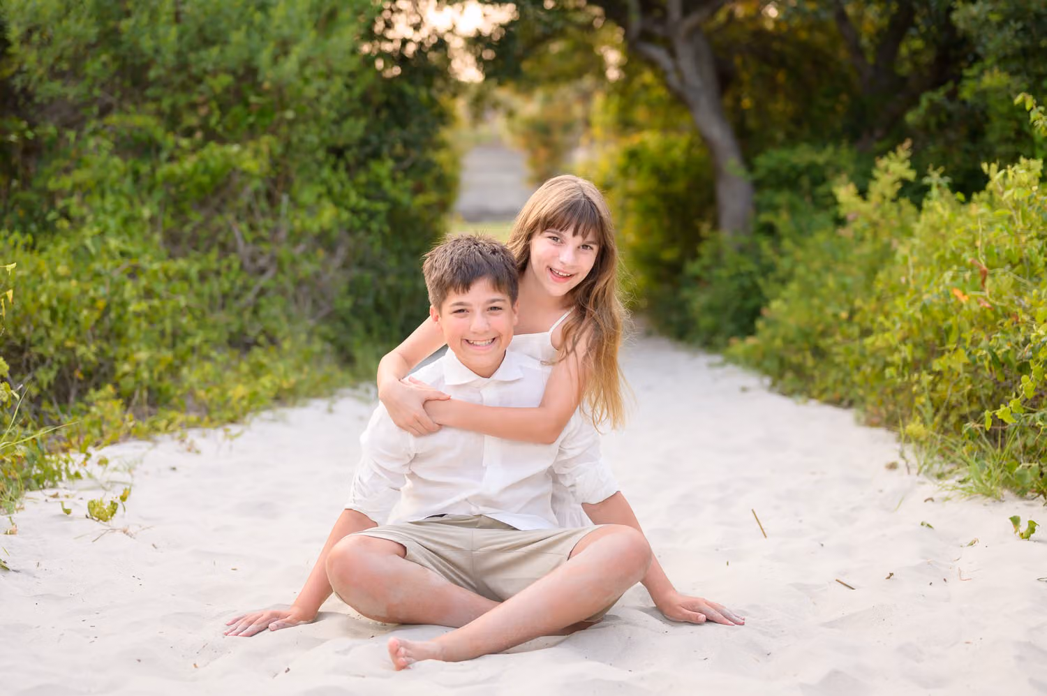 Big sister hugging her little brother from behind as they sit on the sandy beach path at Huntington Beach State Park