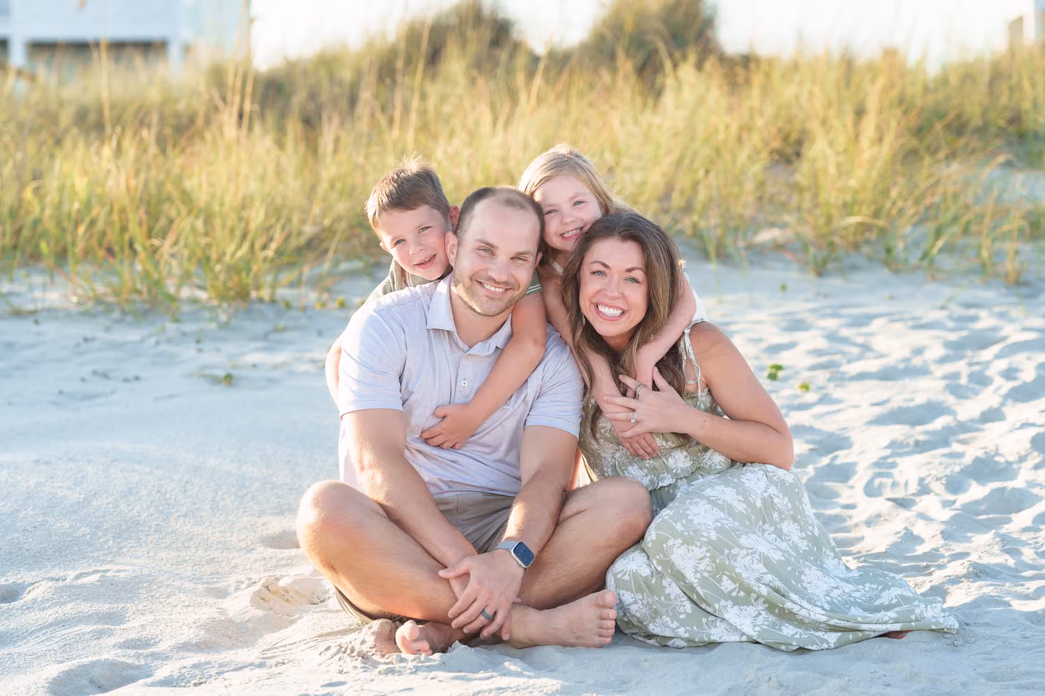 Two kids hugging mom and dad from behind as the parents sit in the sand by the dunes in North Myrtle Beach
