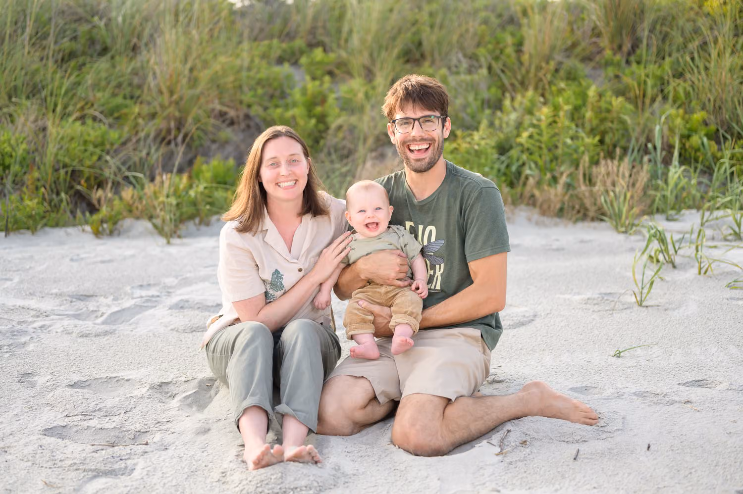 Mom and dad sitting in the sand by the dunes holding their smiling baby boy in North Myrtle Beach