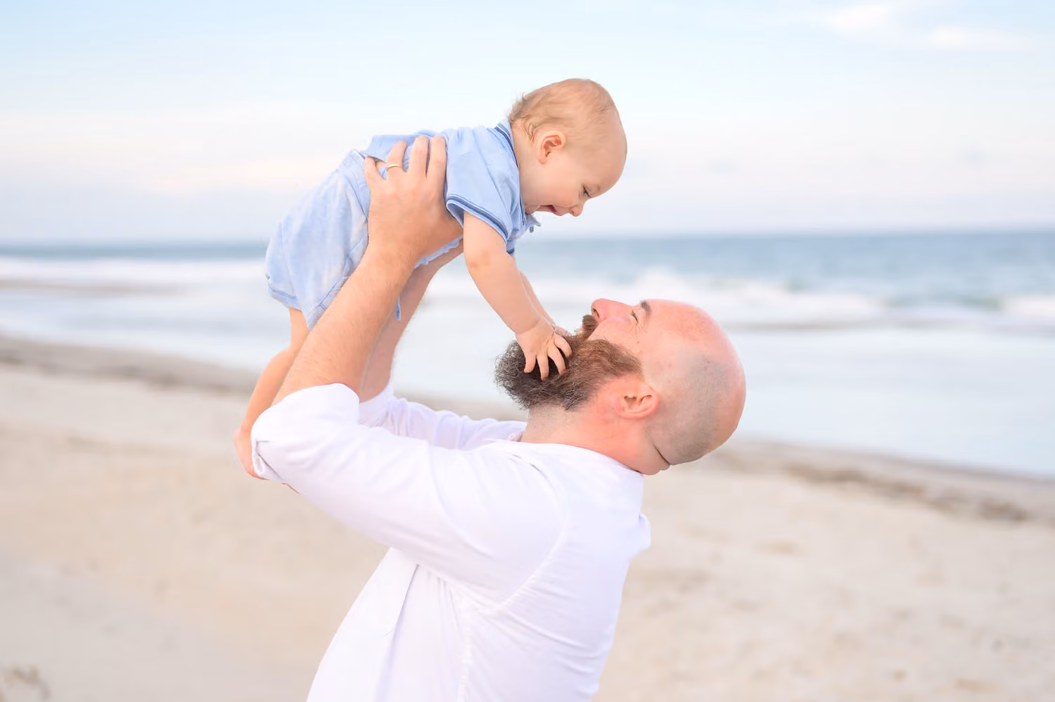 Dad holding his baby boy overhead as the baby grabs his beard on the beach in Myrtle Beach