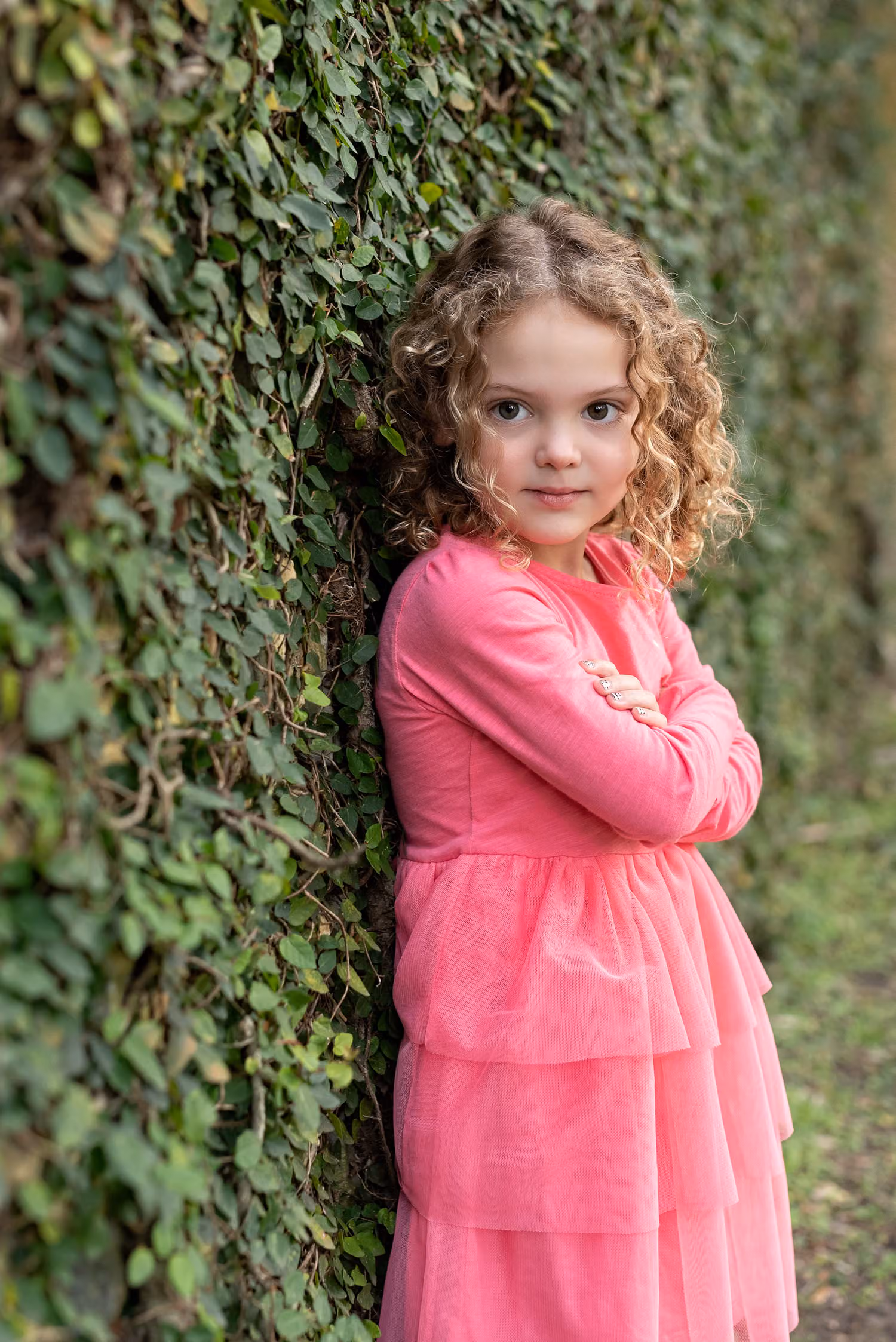 Curly-haired little girl in a pink dress with arms crossed against an ivy-covered wall at Huntington Beach State Park