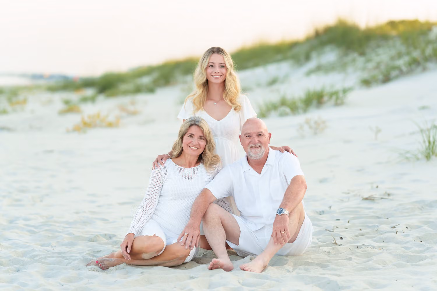 Parents in white sitting in the sand by the dunes with their grown daughter behind them at Huntington Beach State Park
