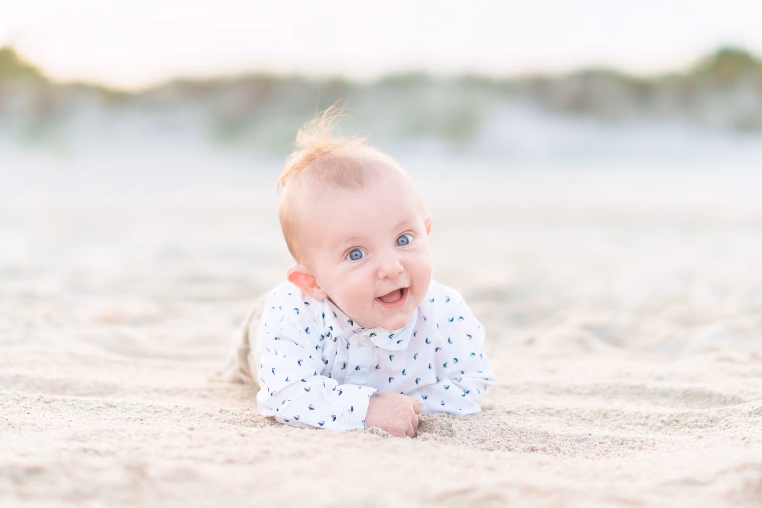 Smiling baby boy in a print shirt lying on his tummy in the sand by the dunes at Huntington Beach State Park