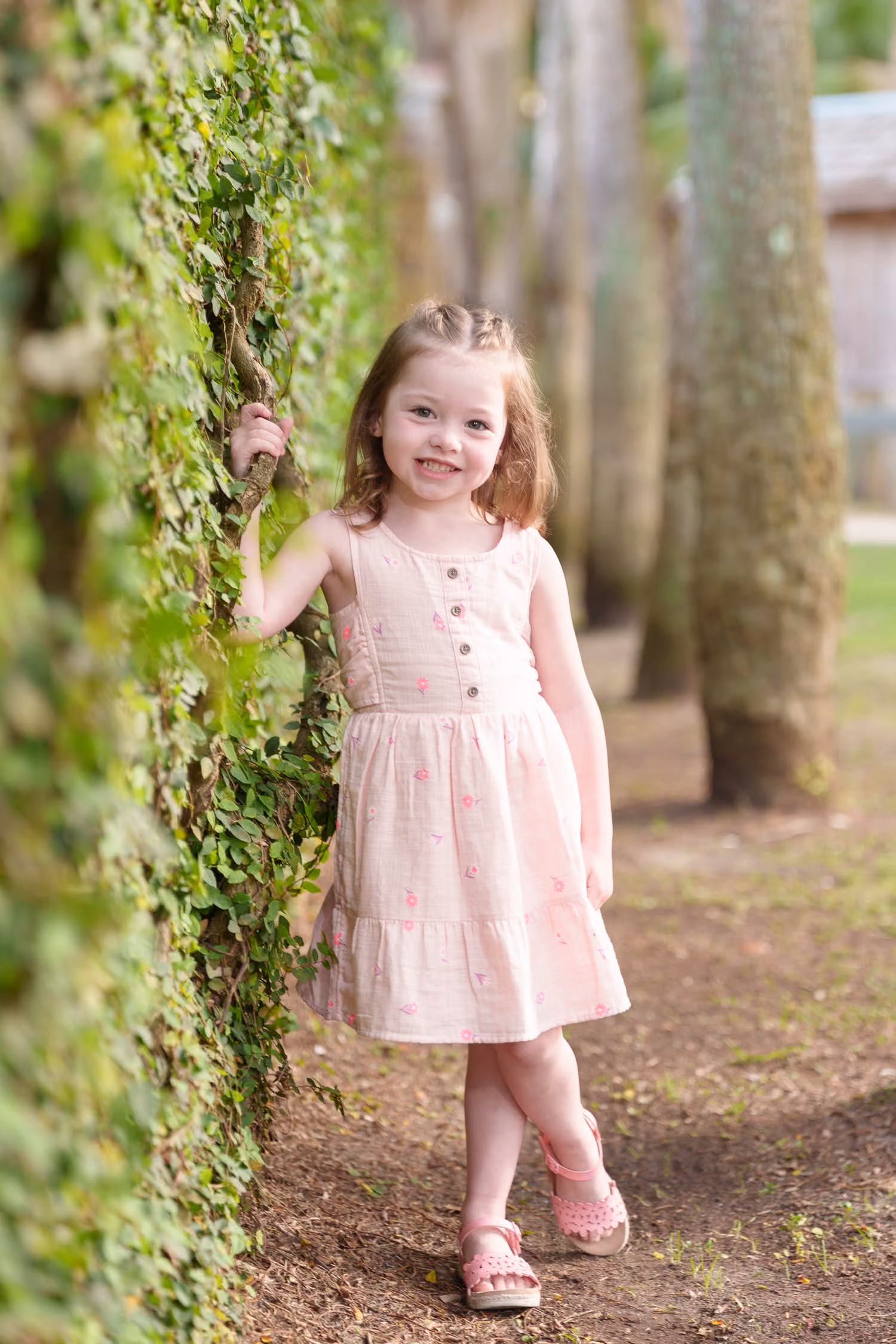 Little girl in a pink dress smiling as she leans against an ivy-covered wall at Huntington Beach State Park