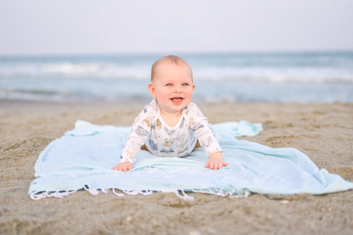 Smiling baby on a light blue blanket on the sand with the ocean behind her in North Myrtle Beach