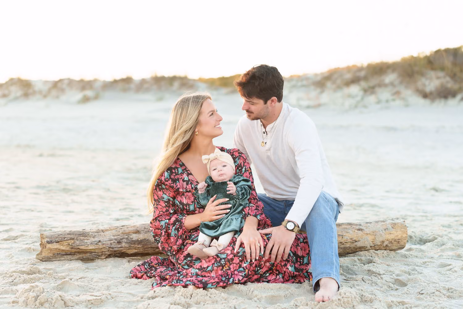 New parents sitting on a driftwood log by the dunes, gazing at their baby girl at Huntington Beach State Park