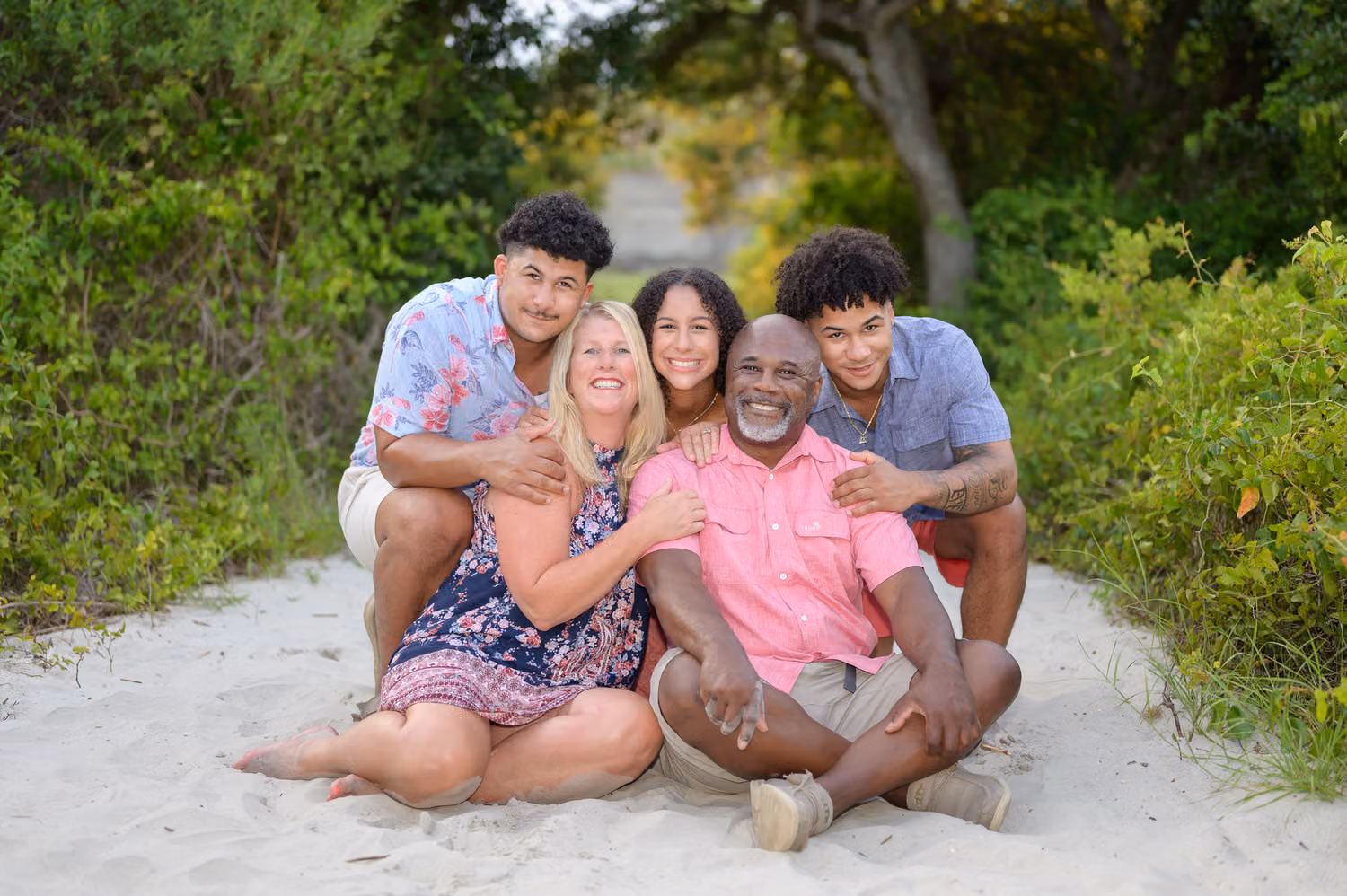 Parents sitting in the sand on the beach path with their three grown kids hugging them at Huntington Beach State Park