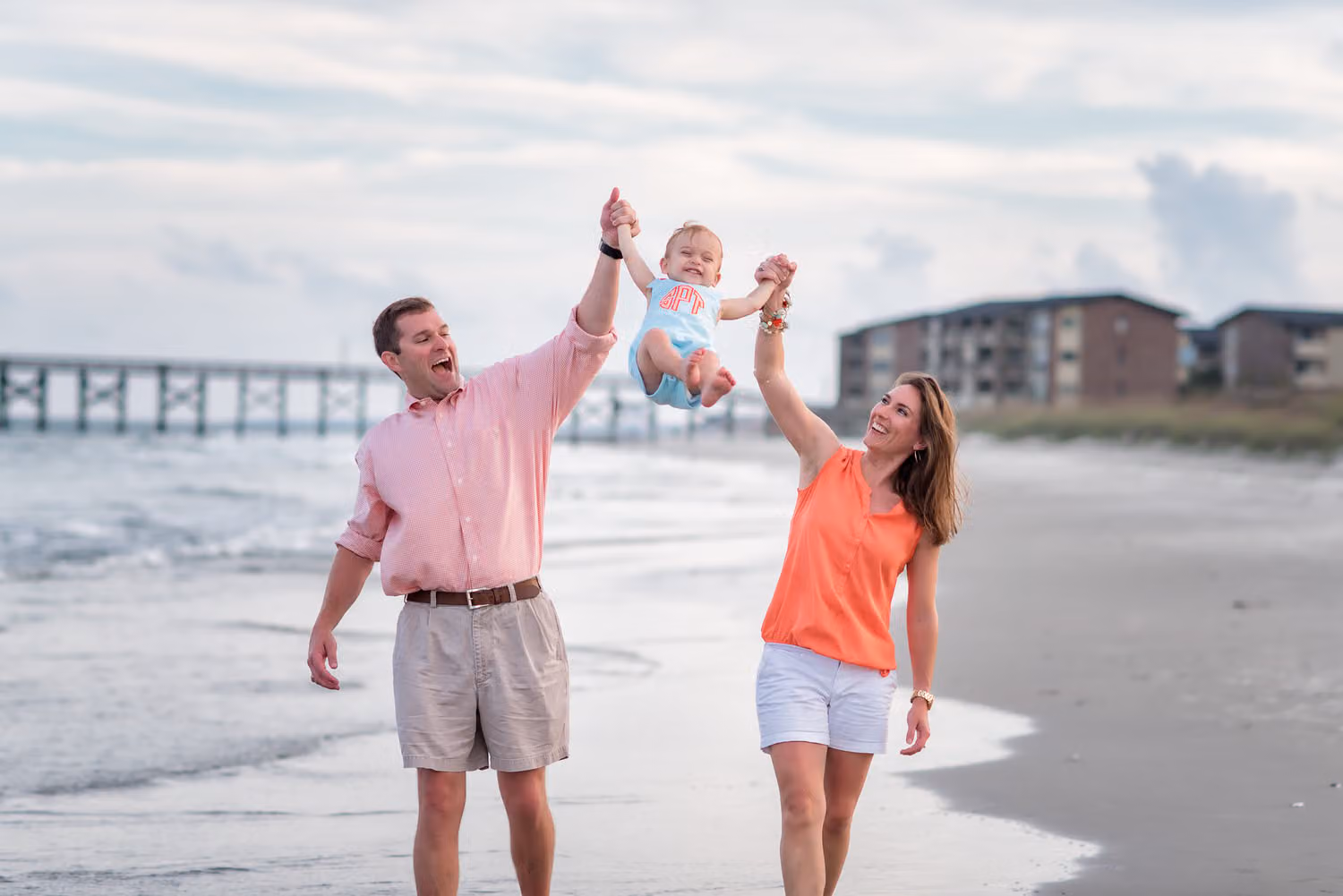Mom and dad swinging their little boy by the arms along the shoreline with a pier behind them in Murrells Inlet
