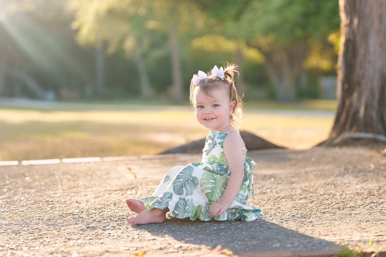 Toddler girl in a floral dress sitting on a path with sun rays shining through the trees at Huntington Beach State Park