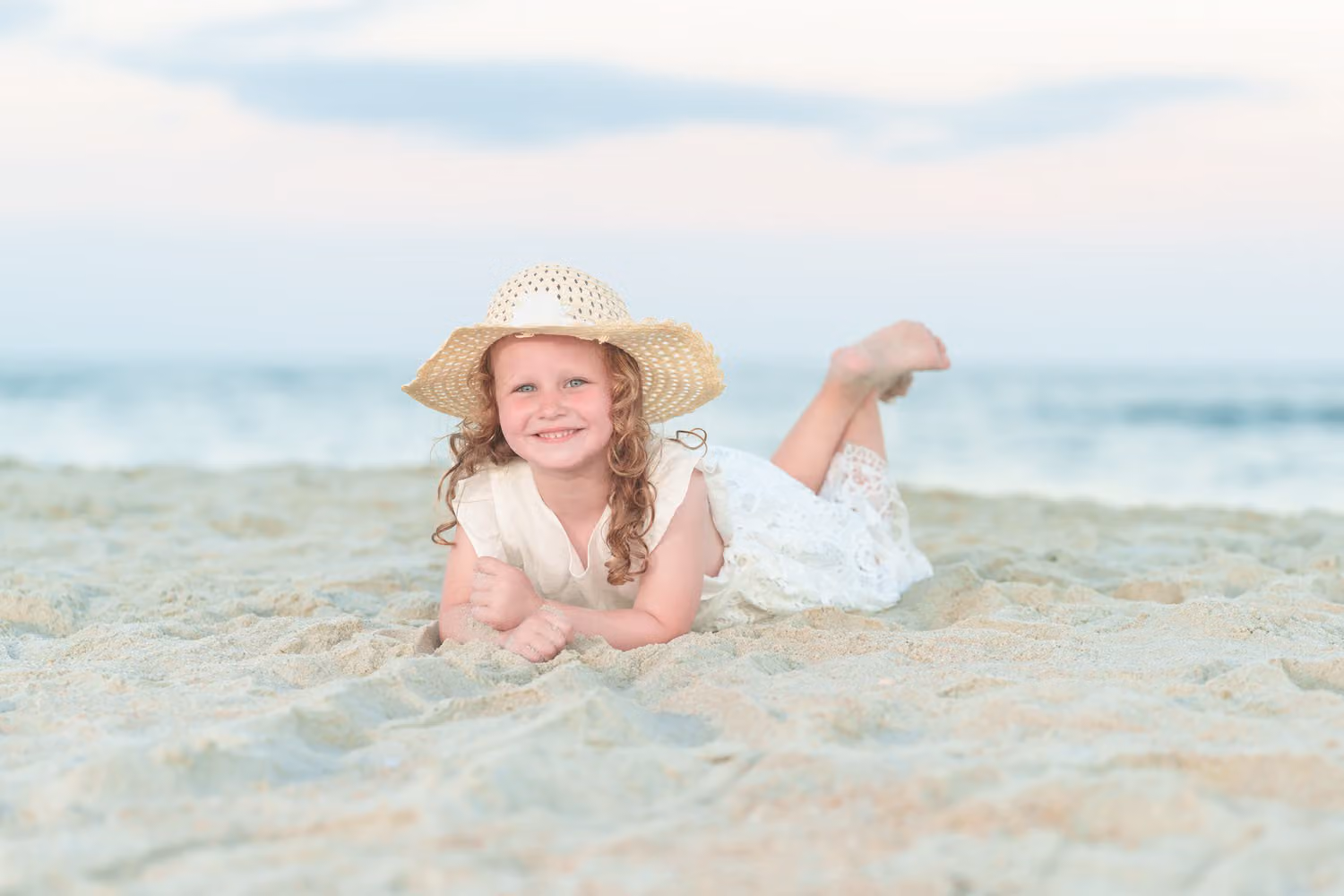 Little girl in a straw hat lying in the sand with her feet kicked up by the ocean at Huntington Beach State Park