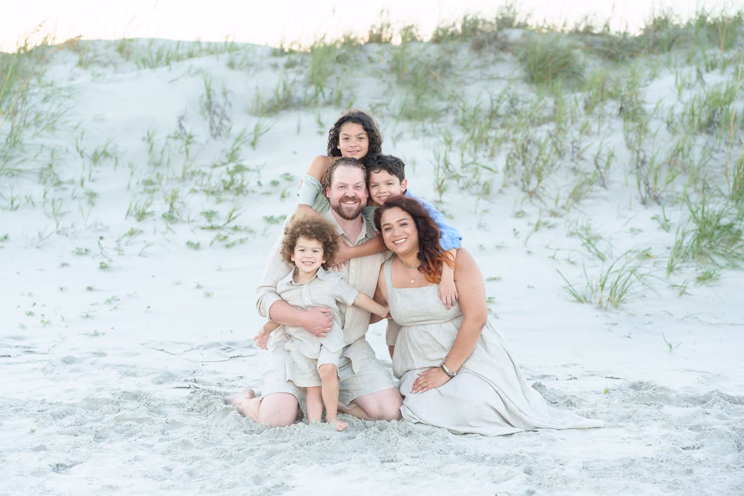 Family of five sitting together in the white sand with dune grass behind them at Huntington Beach State Park