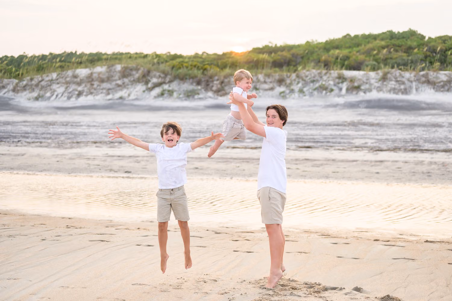 Brothers leaping on the sand while the oldest lifts the baby overhead with the dunes behind at Huntington Beach State Park