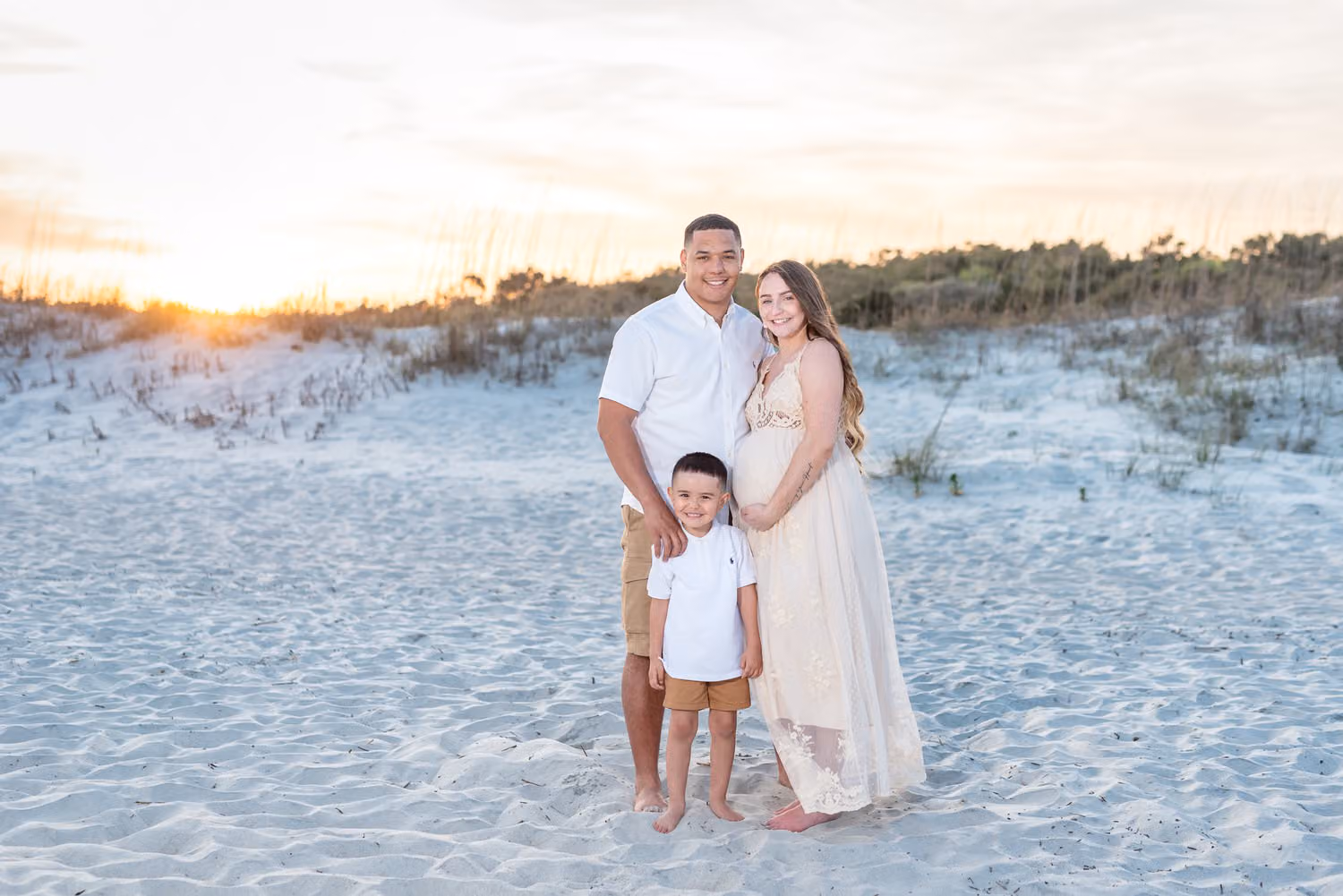 Expectant mom and dad standing with their little boy in front of the dunes at sunset at Huntington Beach State Park