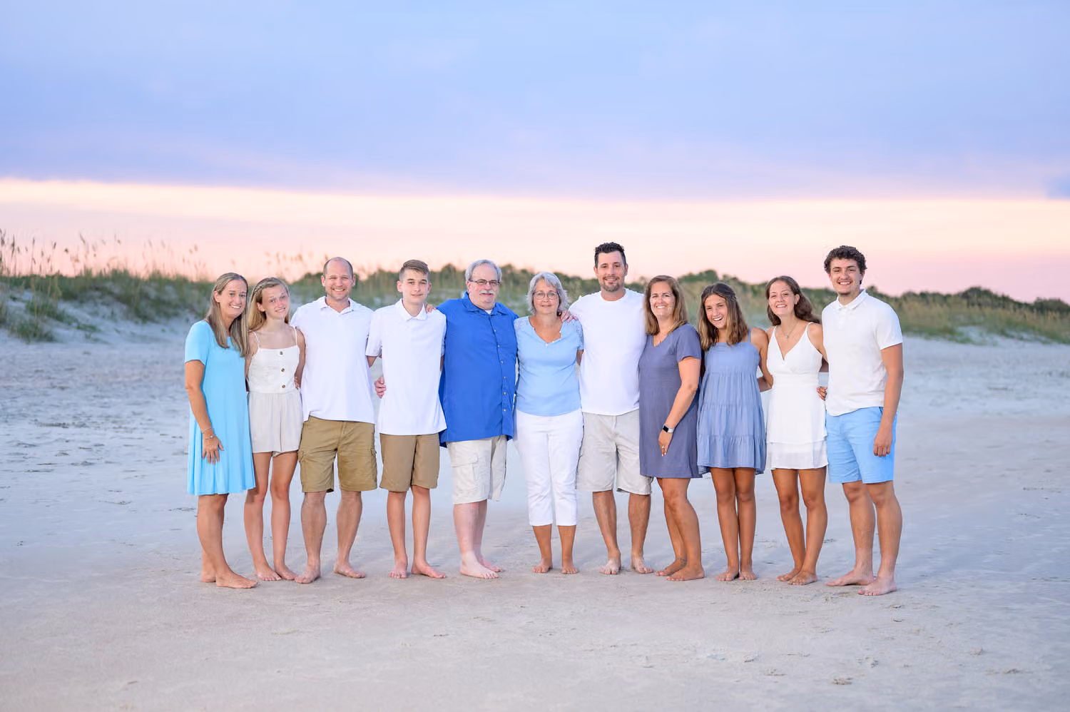 Family of eleven standing in a row in front of the dunes under a pink sunset sky at Huntington Beach State Park