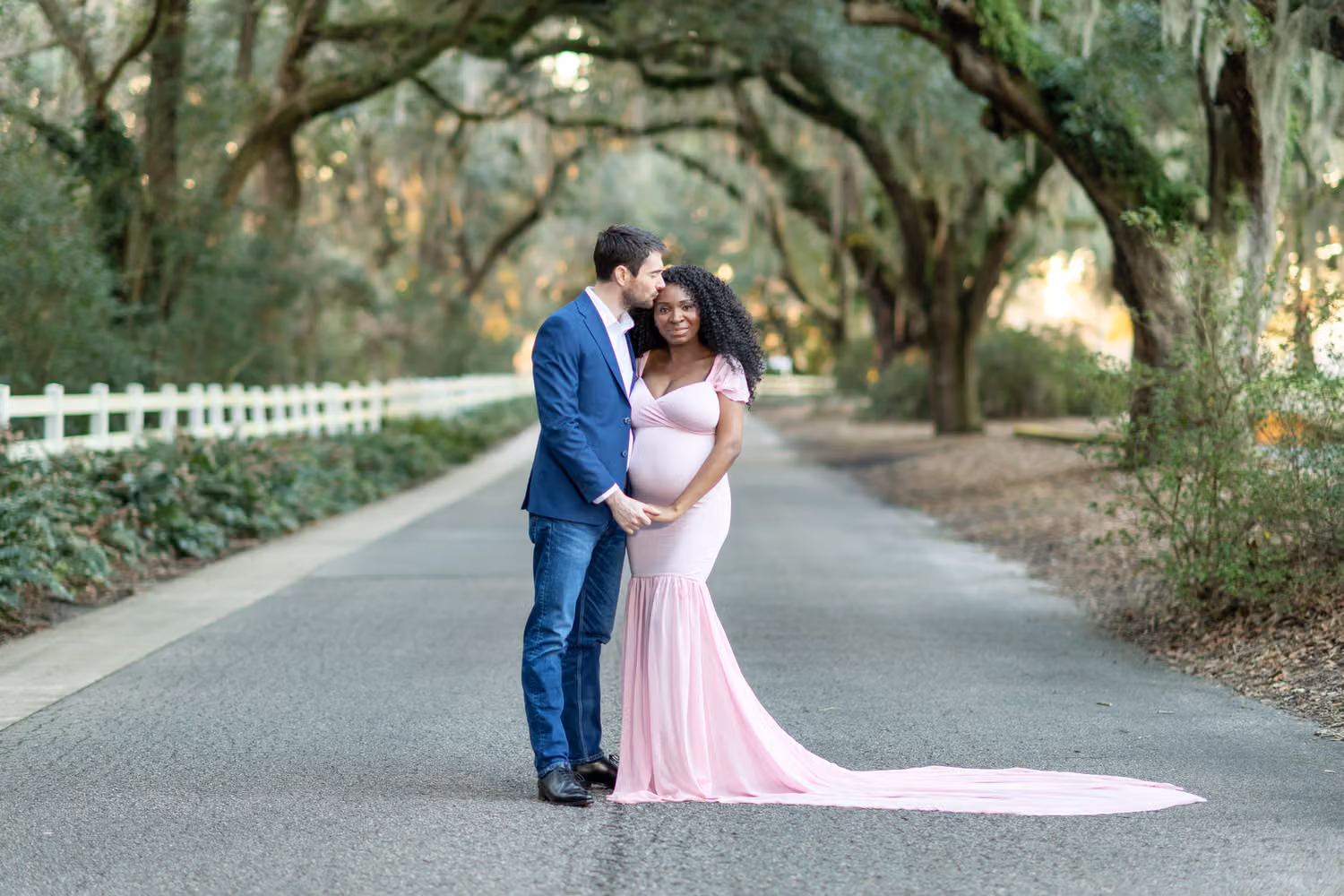 Man kissing his pregnant wife's cheek as she wears a long pink gown on a road under mossy oaks at Caledonia Golf & Fish Club