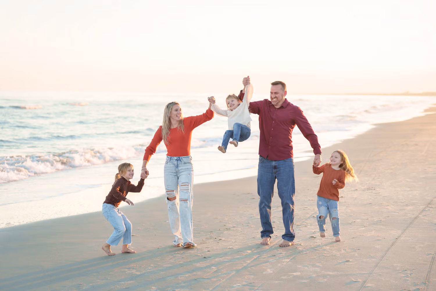 Mom and dad swinging their toddler between them as two big sisters run alongside on the beach at Huntington Beach State Park