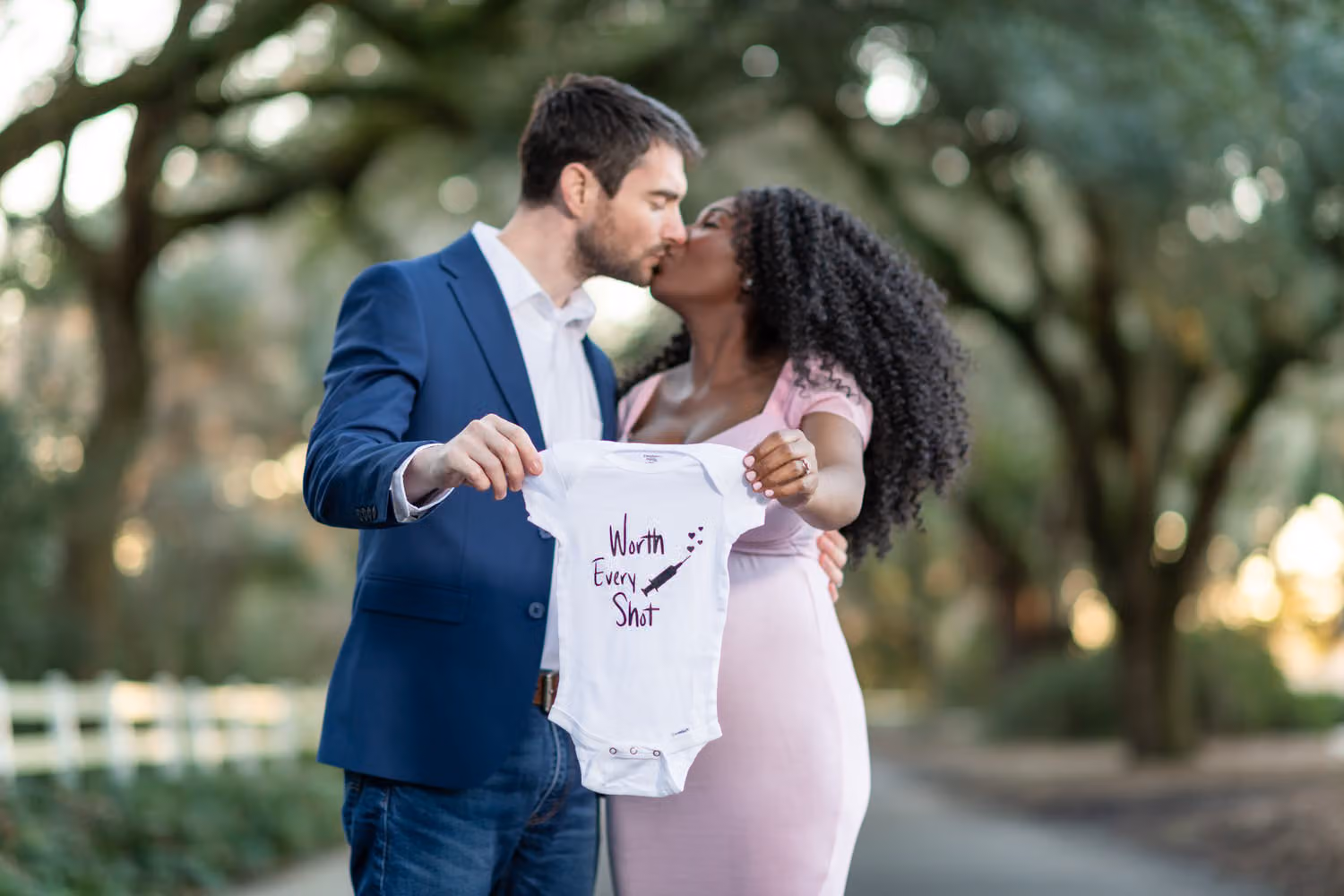 Expectant couple kissing under the oaks while holding a baby onesie that reads Worth Every Shot at Caledonia Golf & Fish Club