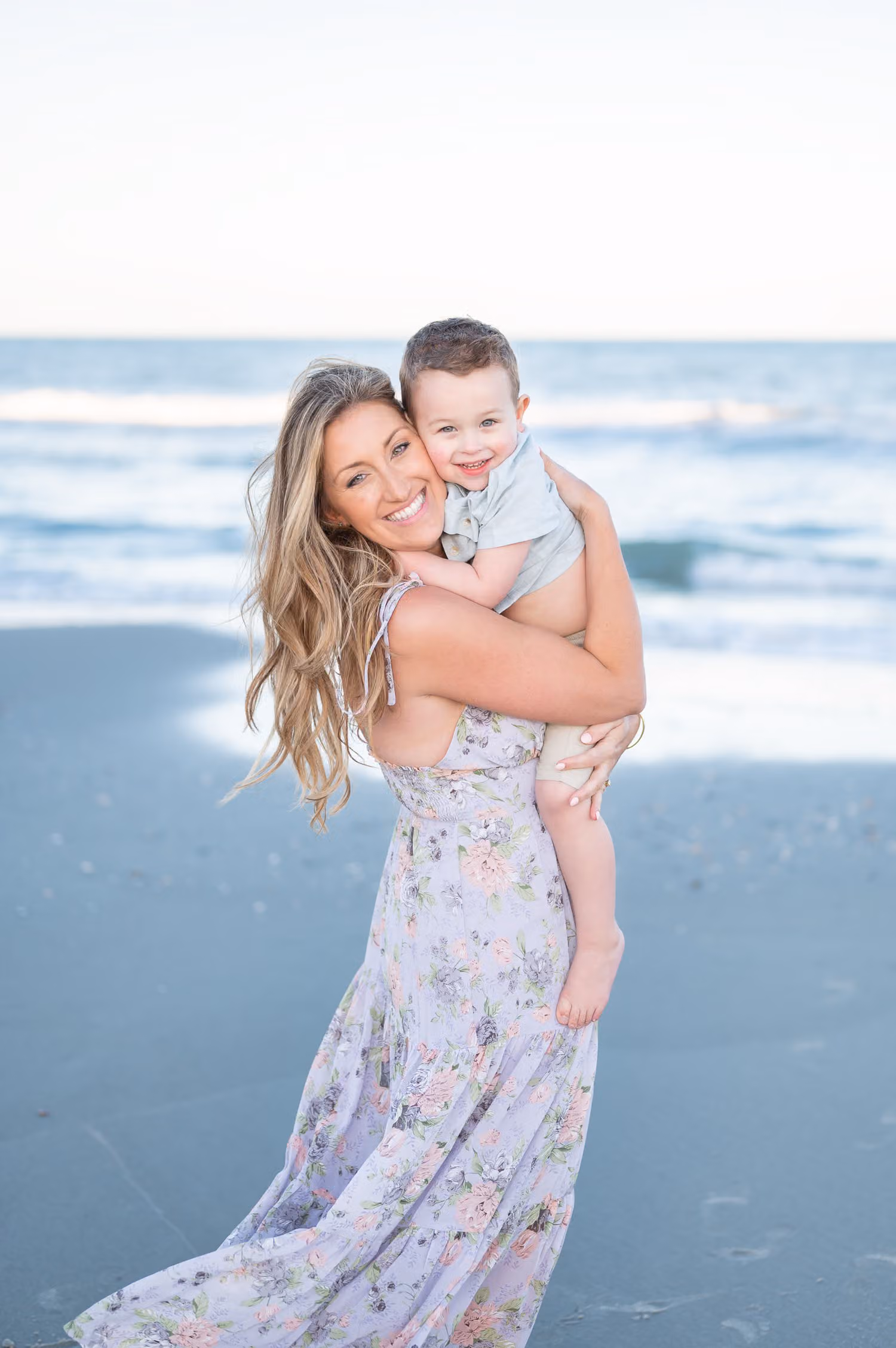 Smiling mom in a floral dress holding her little boy up on her shoulder by the ocean at Garden City Beach