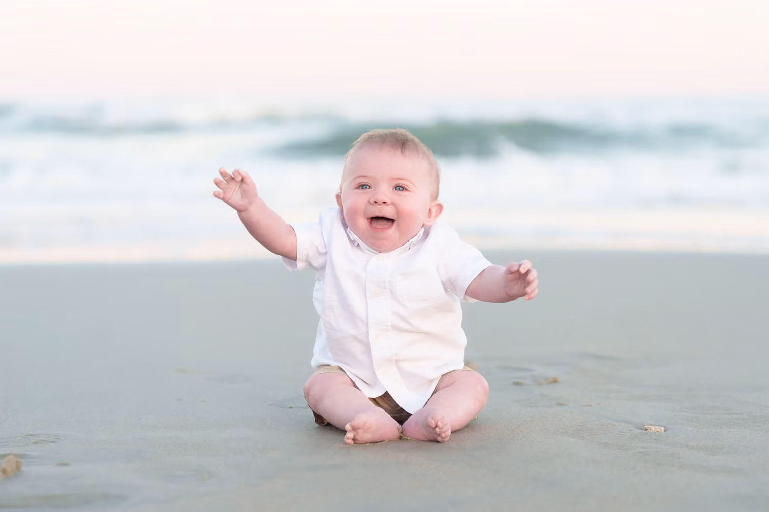 Baby boy in white laughing with his arms out while sitting on wet sand at Huntington Beach State Park