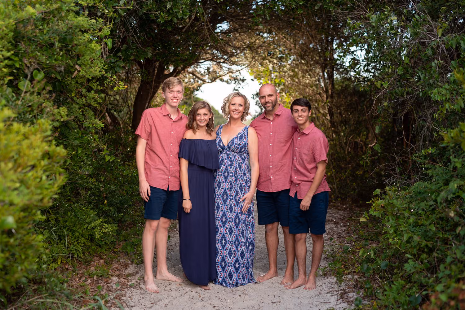 Family of five in coral and navy standing in a tunnel of trees on the path to the beach at Huntington Beach State Park