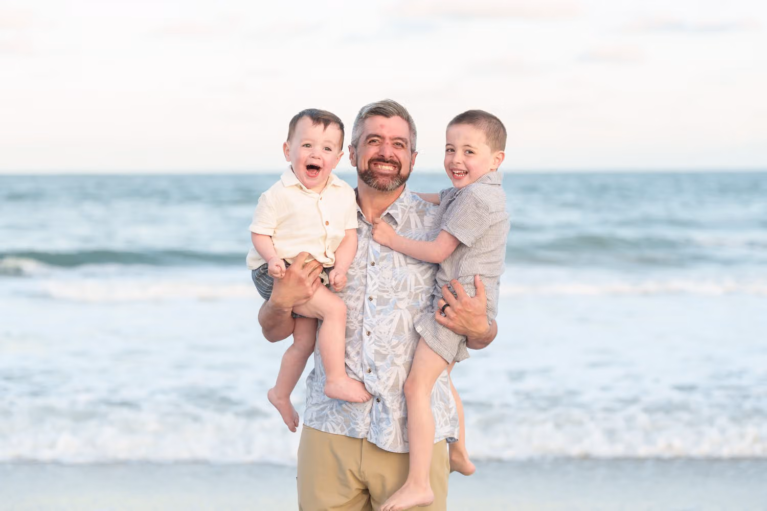 Laughing dad holding one son on his hip while his other son hugs him by the ocean at Huntington Beach State Park