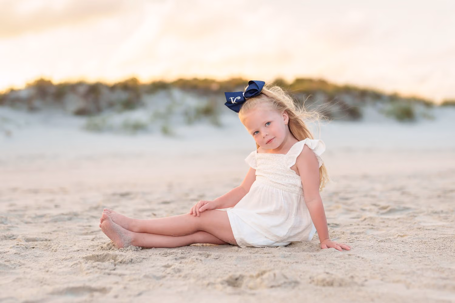 Little girl with a big navy bow sitting in the sand in front of the dunes in warm sunset light at Huntington Beach State Park