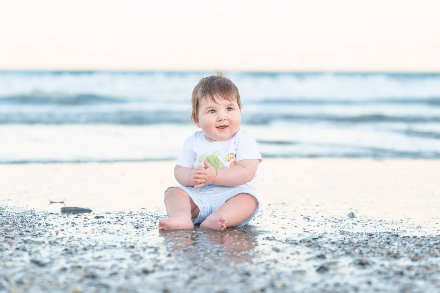 Baby boy in white sitting at the shoreline as the tide washes over the sand in North Myrtle Beach