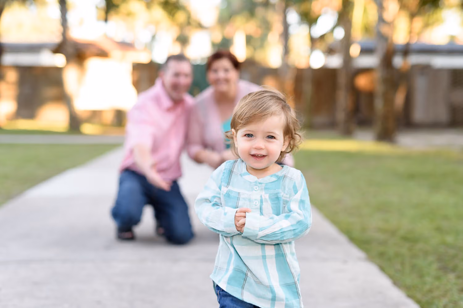 Toddler girl walking toward the camera on a path while her parents kneel behind her at Huntington Beach State Park