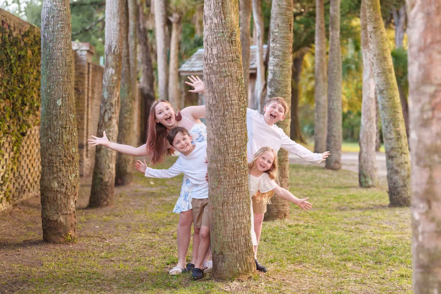 Brothers and sisters playfully leaning out from behind a row of palm tree trunks at Huntington Beach State Park