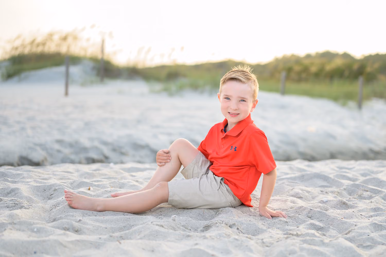 Little boy in a red polo sitting back in the sand in front of the dunes at Myrtle Beach State Park