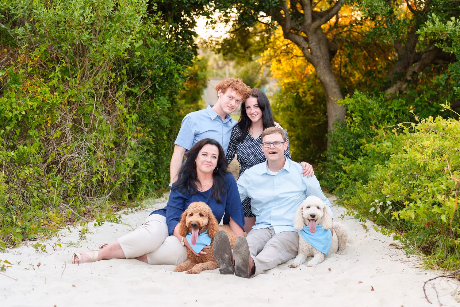 Family of four sitting on a sandy path under the trees with their two Goldendoodles in front of them